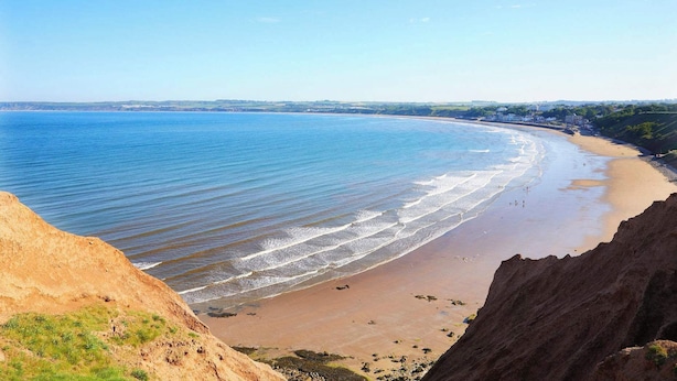 Une plage côtière aux eaux d’un bleu limpide, avec un rivage sablonneux et une falaise surplombant la plage.