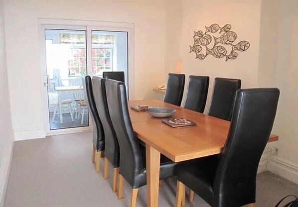 A dining area with a wooden table and black chairs, a decorative wall piece, and a view through a glass door. - Ivy House - Seafront house with garden seating and stunning sea views! (Filey)