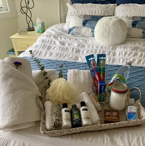 Un lit bien fait, recouvert d’une couverture à rayures bleues et blanches, avec un oreiller blanc et un coussin décoratif. Une tablette contenant des articles de toilette, notamment des brosses à dents, un flacon de gel hydroalcoolique et un pot de miel, est posée sur le lit.