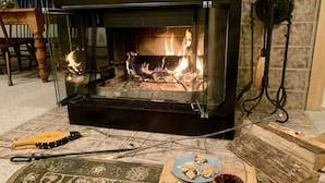 A fireplace with visible logs and flames, a wooden table with a plate of cookies, a bowl of marshmallows, and a glass of red wine. - Summer Escape Near Lakes, Trails & Mt Snow (Dover)