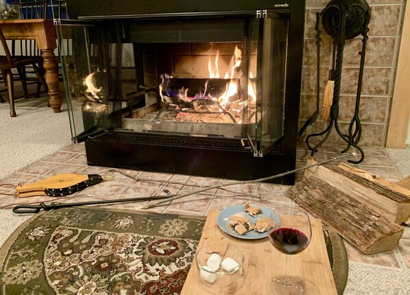 A fireplace with visible logs and flames, a wooden table with a plate of cookies, a bowl of marshmallows, and a glass of red wine.
