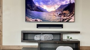 Un televisor de pantalla plana montado en la pared sobre un mueble bajo de madera con estantes y un otoman blanco.