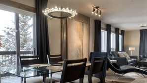 Una sala de estar moderna con una mesa de comedor de vidrio, sillas de madera oscura, un sofá y una mesa de centro. Hay una ventana grande con cortinas, un candelabro y vistas a árboles cubiertos de nieve.