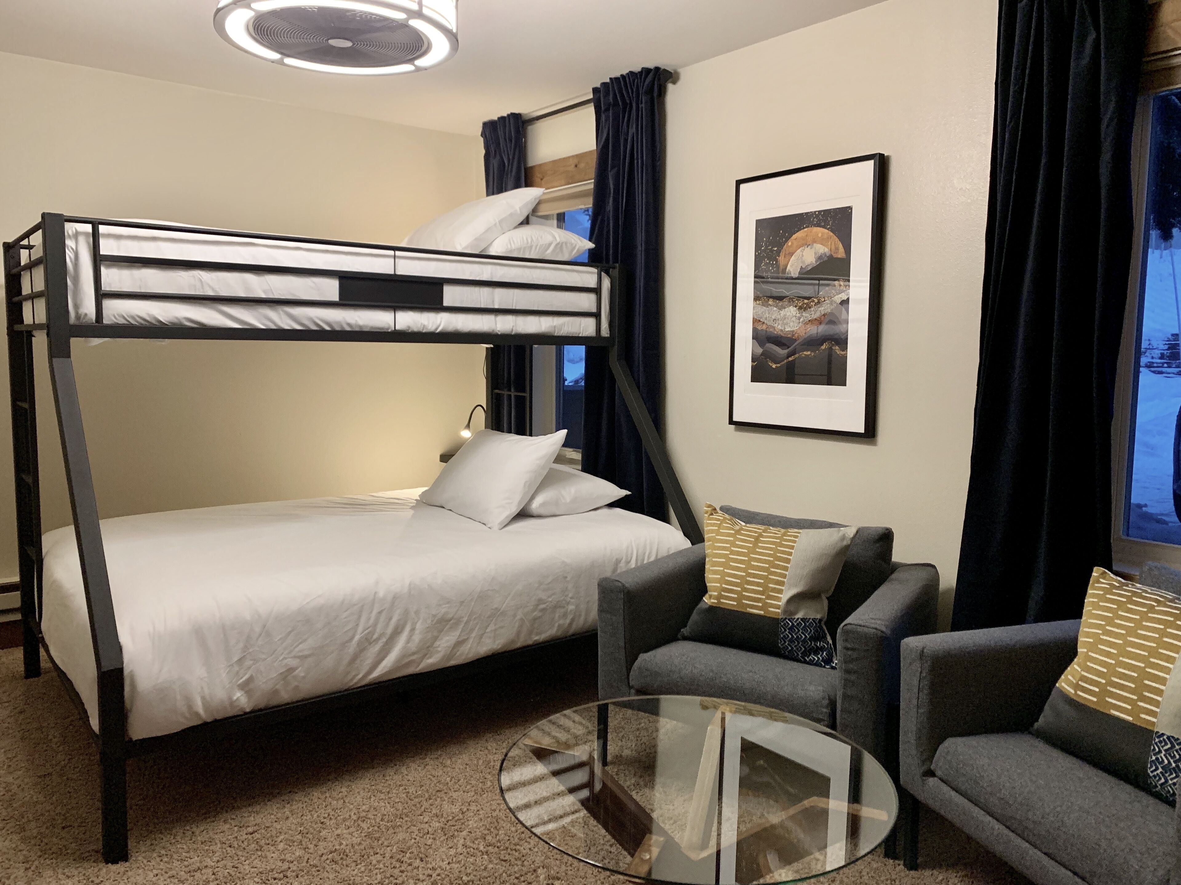 Una cama litera con ropa de cama blanca, una mesita de centro de cristal, dos sillones grises y un cuadro en la pared.
