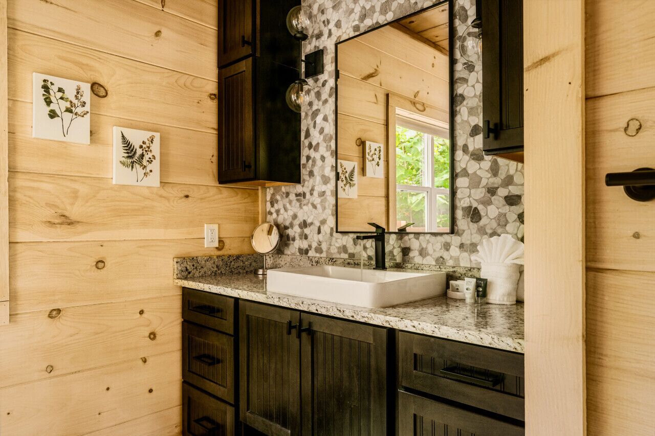 Un bagno con pareti in legno, un piano di lavoro in granito, un lavabo bianco, mobili scuri e uno specchio con zoccolo in pietra.
