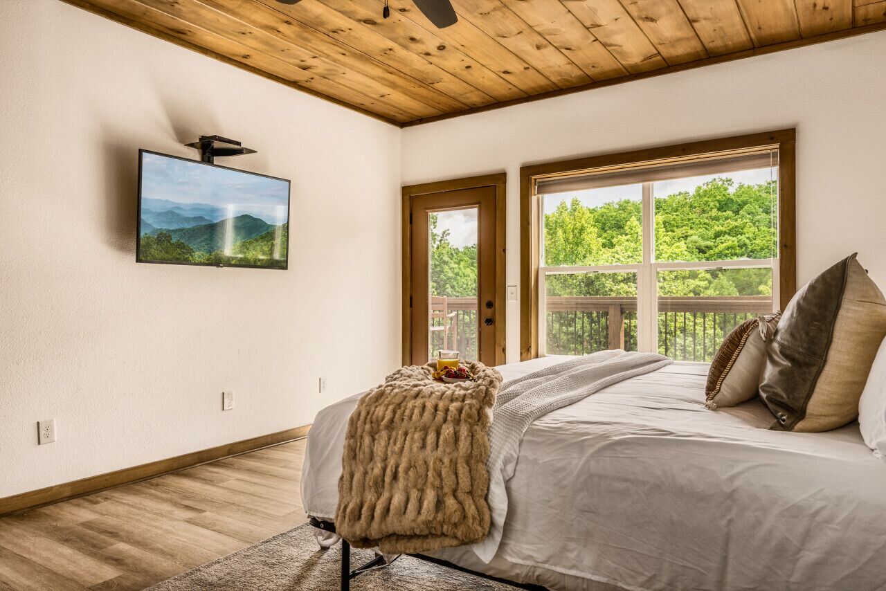 Un dormitorio con una cama grande, televisor montado, techo de madera y ventana con vista a la vegetación.