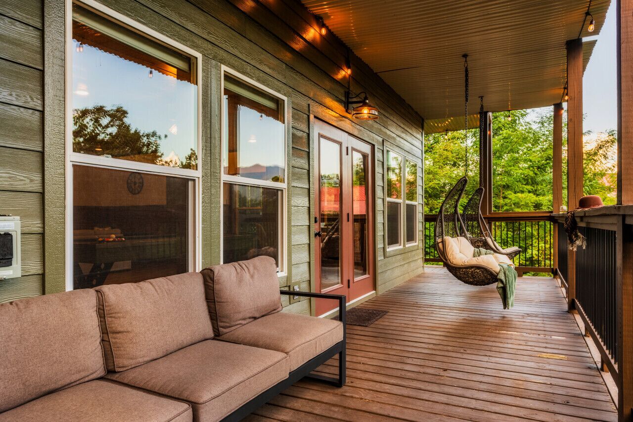 Une terrasse en bois avec un canapé et des fauteuils suspendus, de grandes fenêtres et un espace couvert avec une porte.