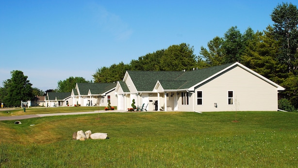 A row of white single-story houses with green roofs, set in a grassy area with trees in the background.