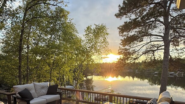 Una terraza de madera con mobiliario exterior, vistas a un lago y árboles al fondo.