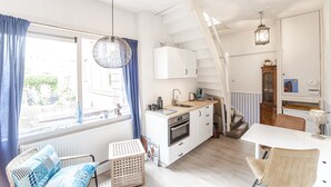 Een kleine keuken met een wit fornuis, een spoelbak en een houten aanrecht. Er staat een stoel en een klein tafeltje bij het raam, dat blauwe gordijnen heeft. Een trap leidt naar een hoger niveau.
