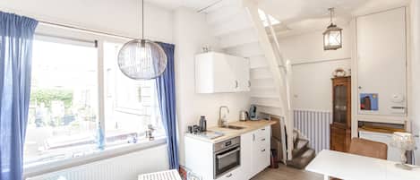Et kompakt køkken med en hvid komfur, en vask og en træbordplade. Der er en stol og et lille bord ved vinduet, som har blå gardiner. En trappe fører til en etage ovenover.