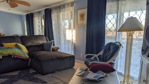 En stue med sofa, lænestol og sofabord. Der er et vindue med gardiner, en lampe og et billede i ramme på væggen.