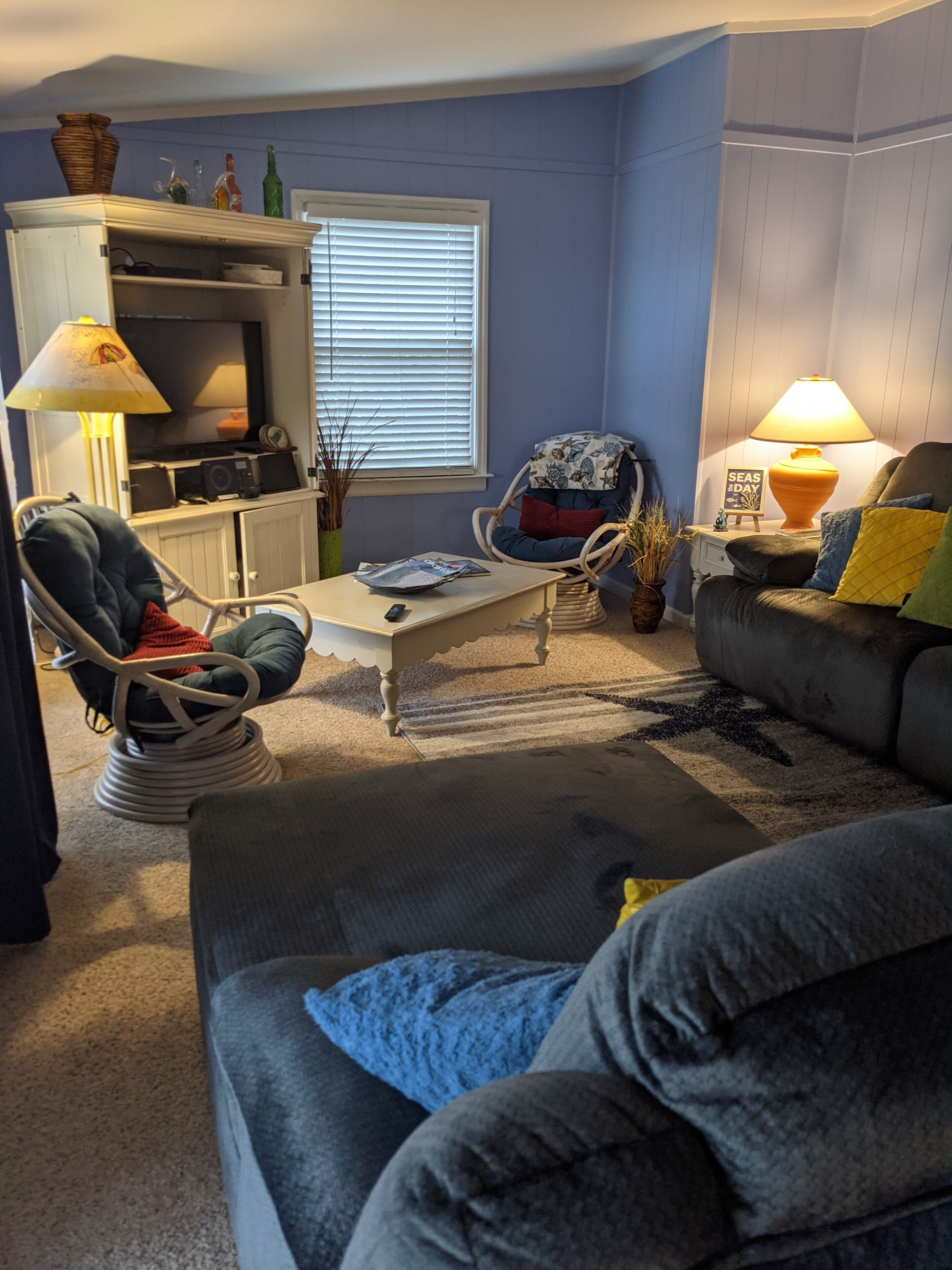 Sala de estar com sofá, poltrona, mesa de centro e uma mesinha de apoio com abajur. Há uma janela com persianas, um armário e um vaso decorativo.
