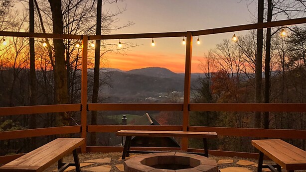 An outdoor seating area with wooden benches and a fire pit, overlooking a scenic view of mountains and a sunset. - Welcome to Bearfeet Retreat, a pristine cabin in a quiet corner in the heart of Wears Valley. (Sevierville)