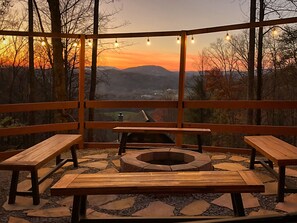 Un espace extérieur aménagé avec des bancs en bois et un foyer, offrant une vue panoramique sur les montagnes et le coucher de soleil.