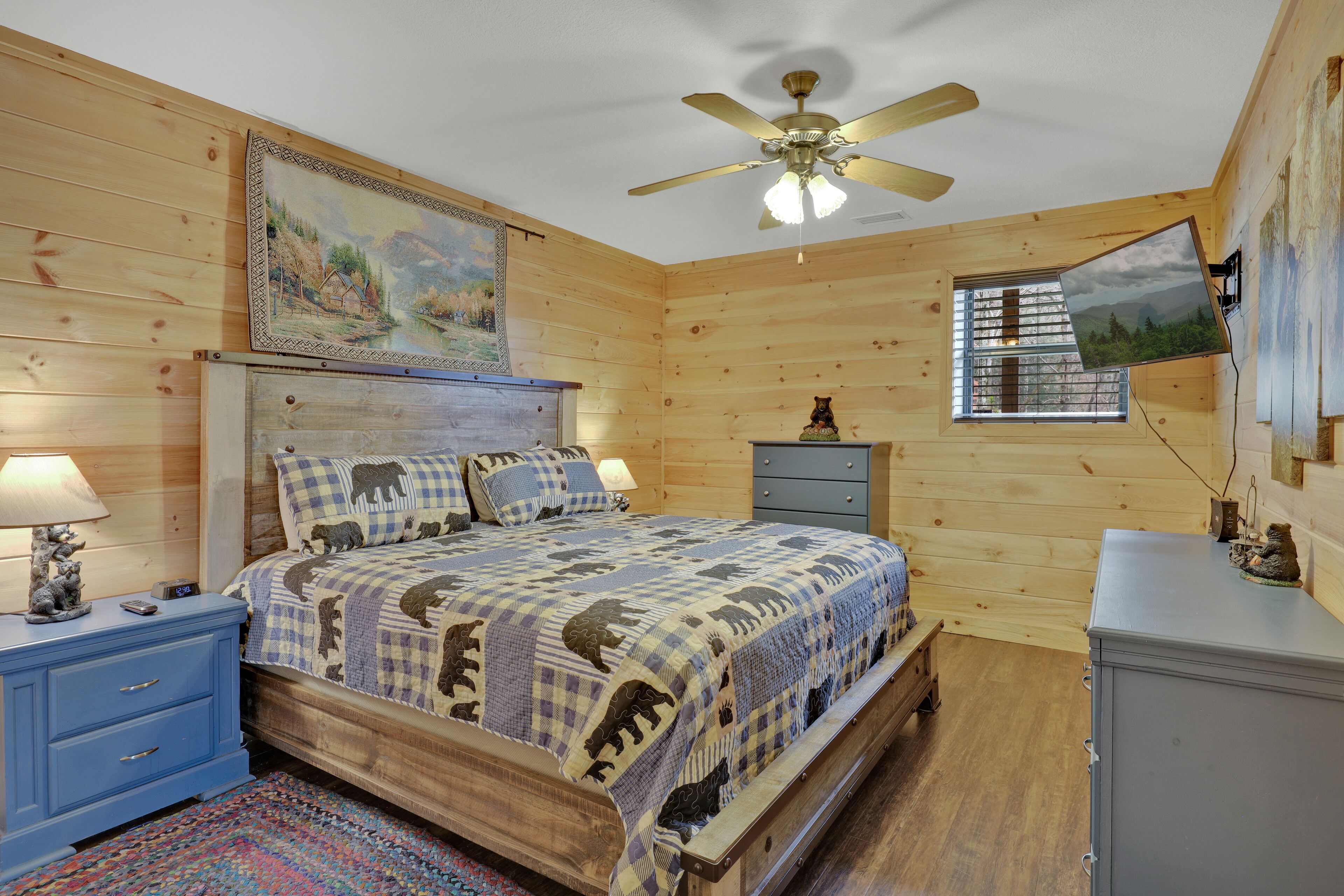 A wooden cabin bedroom with a bed, a nightstand, a dresser, a ceiling fan, and a window with a view of trees.