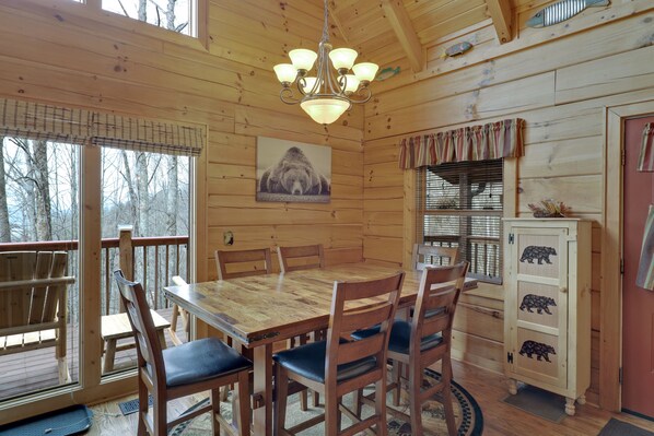 Une salle à manger en bois, avec une table et des chaises, un lustre, un meuble décoré sur le thème de l’ours et une fenêtre avec des stores.