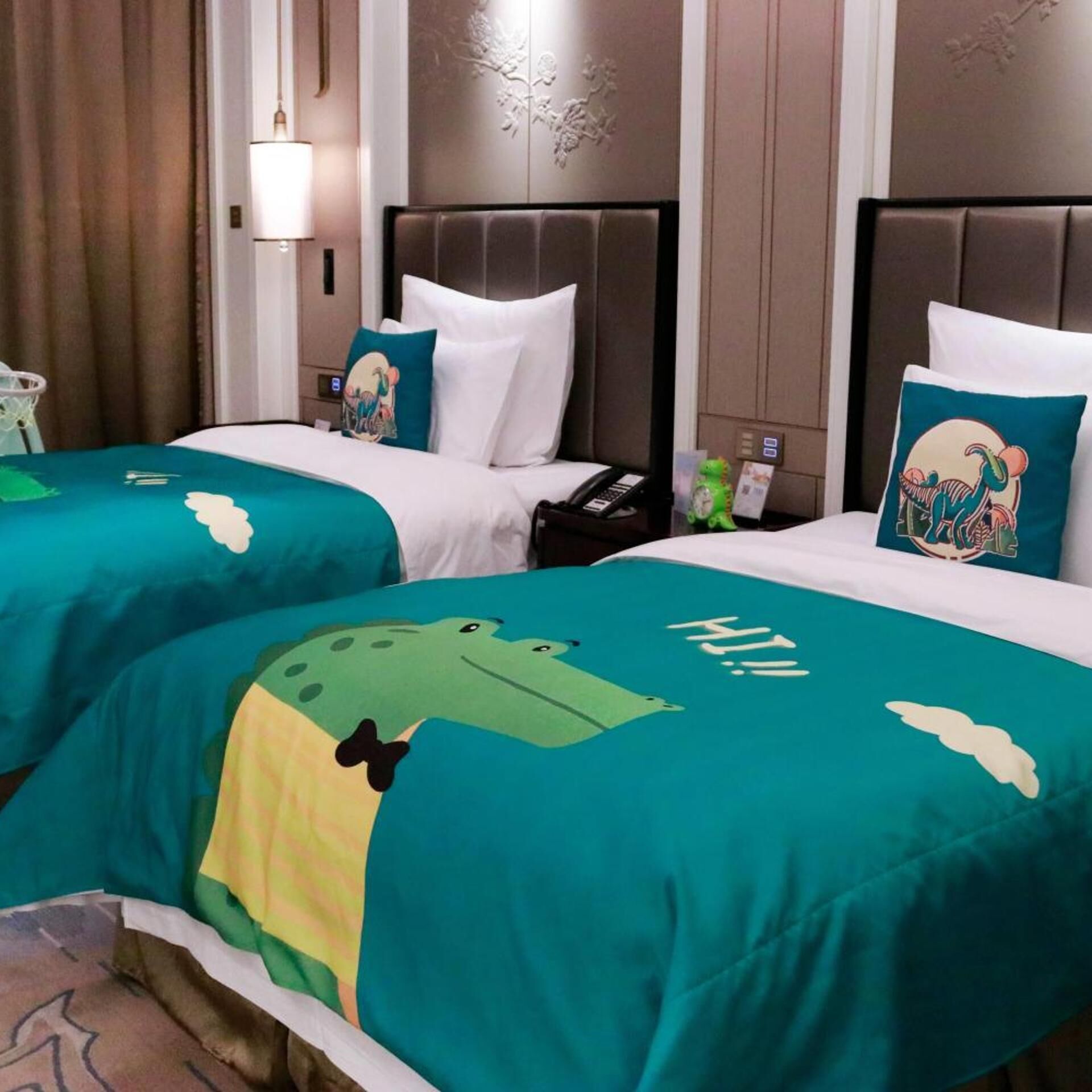 Two hotel beds with teal bedspreads featuring a cartoon frog design and the word 'HI!' on the bedspread.
