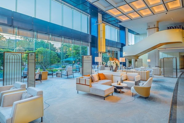 Un hall espacioso con un diseño moderno, que cuenta con una amplia zona de estar con sofás y sillones, una escalera y grandes ventanales con vistas al exterior.