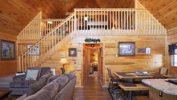A log cabin interior with a staircase leading to a loft, a living area with a sofa, a dining table, and a kitchen area. - Cabin w/ loft & beautiful updates, indoor fun/hot tub/new grill & communal pool! (Sevierville)