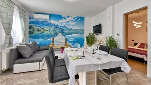 A dining area with a table set for two, a grey sofa, a TV mounted on the wall, and a large scenic mural of a lake and mountains. - Nice home in Rivalno with sauna (Rivalno)
