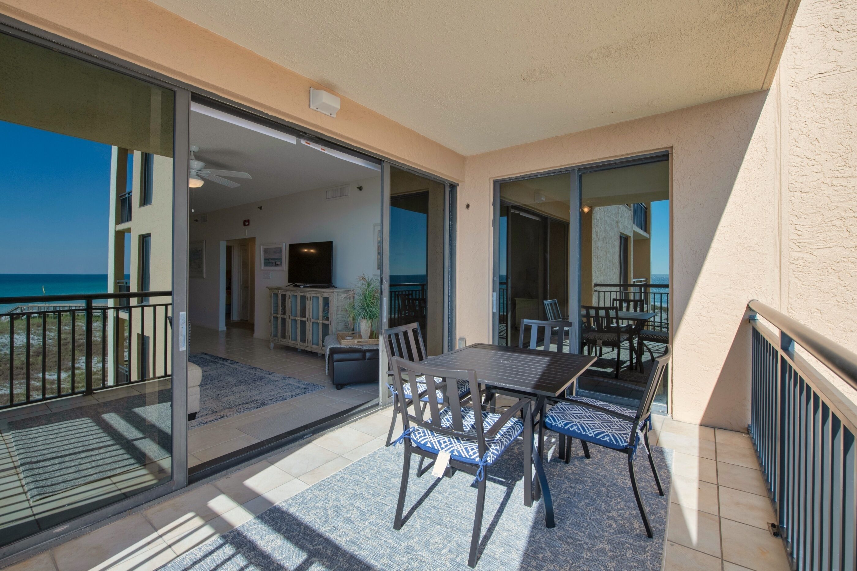 Varanda com mesa e cadeiras, porta de vidro deslizante que dá para um quarto com TV, e vista para o oceano.