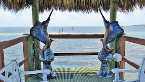 Une structure à toit de chaume, agrémentée de deux sculptures de poissons en métal, se dresse sur une terrasse en bois offrant une vue sur la mer.