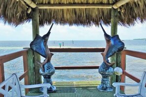 Estructura con techo de paja y dos esculturas de peces metálicos en una terraza de madera con vista al mar.