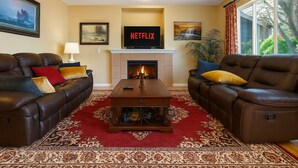 A living room with two leather sofas, a fireplace, a Netflix sign, and a large window with a view of trees. - Experience Seattle Hot Tub Firepit Backyard BBQ EV2 King Bed (Lynnwood)