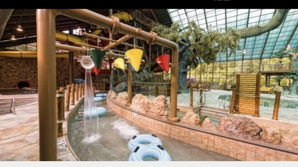 Un espace de jeux aquatiques intérieur doté d’un plafond en verre, de formations rocheuses et d’éléments décoratifs colorés suspendus.