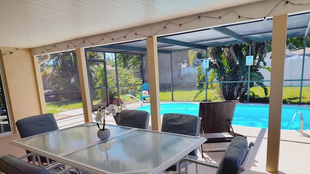Un patio couvert avec une table et des chaises en verre, un petit vase blanc orné de fleurs et une vue sur une piscine et la verdure environnante.
