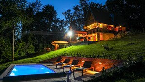 Una cabaña de dos pisos con piscina, mobiliario exterior e iluminación destacada en la fachada.