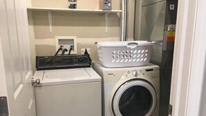 Un área de lavandería compacta con lavadora y secadora de carga frontal, una cesta sobre la secadora y un estante superior con un recipiente y un pequeño electrodoméstico.