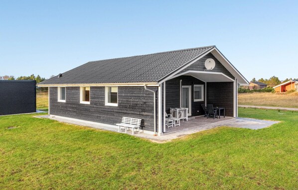 A small, single-story building with a dark exterior, a pitched roof, and a covered porch with a bench and chairs. - 2 bedroom lovely home in Skjern (Skjern)