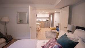 Ein modernes Schlafzimmer mit einem großen Bett, einem Nachttisch und einem Beistelltisch. Das Zimmer hat eine Tür zu einem Küchenbereich mit Kühlschrank, Herd und Esstisch.