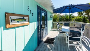 A turquoise building with a door marked "317", a framed fish painting, and a patio with a table and chairs under a blue umbrella.