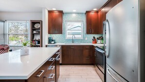 A modern kitchen with a large stainless steel refrigerator, wooden cabinets, a white countertop, and a window with a view of a house.