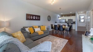 Ein modernes Wohnzimmer mit einem grauen Sofa, gelben Kissen, einer Essecke mit Tisch und Stühlen sowie einer Küche im Hintergrund.