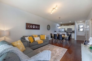 A modern living room with a grey sofa, yellow cushions, a dining area with a table and chairs, and a kitchen in the background.