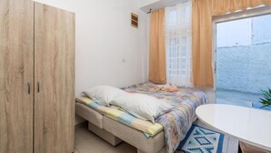 A small, well-lit room with a bed, a wooden wardrobe, a small table, and a window with curtains.