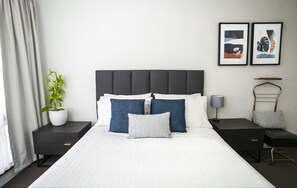 A modern hotel room with a large bed, two bedside tables, a bedside lamp, a potted plant, and two framed artworks on the wall.