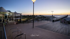 A waterfront promenade with a wooden deck, a street lamp, and a covered seating area with tables and chairs. - View One Three (Seaton)