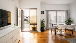A modern living room with a flat-screen TV, a dining area with white chairs, and a sliding glass door leading to a balcony. - View One Three (Seaton)