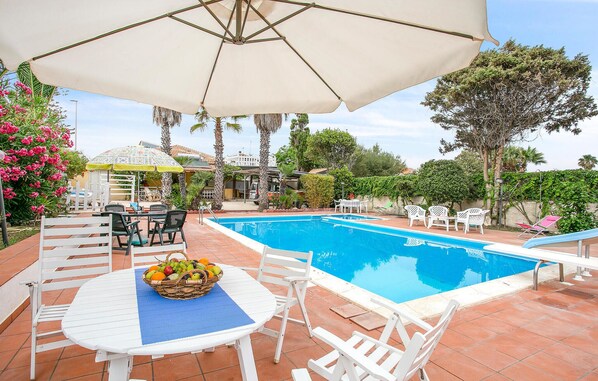 Piscina com cadeiras de descanso brancas e uma mesa com cesta de frutas.