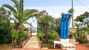 Een tuin met een pad dat naar een strand leidt, met palmbomen, roze bloemen en een blauw beeldhouwwerk.
