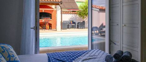 A hotel room with a bed, a view of a swimming pool, and a barbecue area.