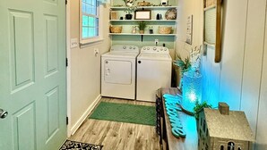 A laundry room with two white washing machines, a small window with a curtain, and a shelf with various items. - Country Escape Home in the Woods- Chesapeake, VA (Chesapeake)