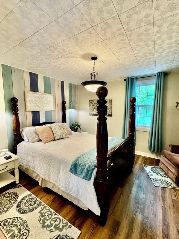 Un dormitorio con cama con dosel, suelo de madera, ventilador de techo, ventana con cortinas y mesita de noche con lámpara.