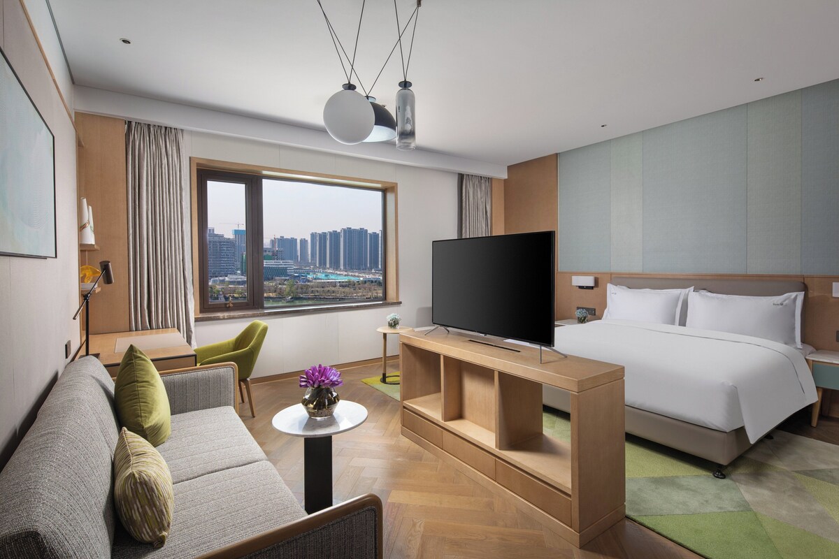 Ein modernes Hotelzimmer mit einem großen Bett, einem Flachbildfernseher, einer Sitzecke und Blick auf die Stadt.