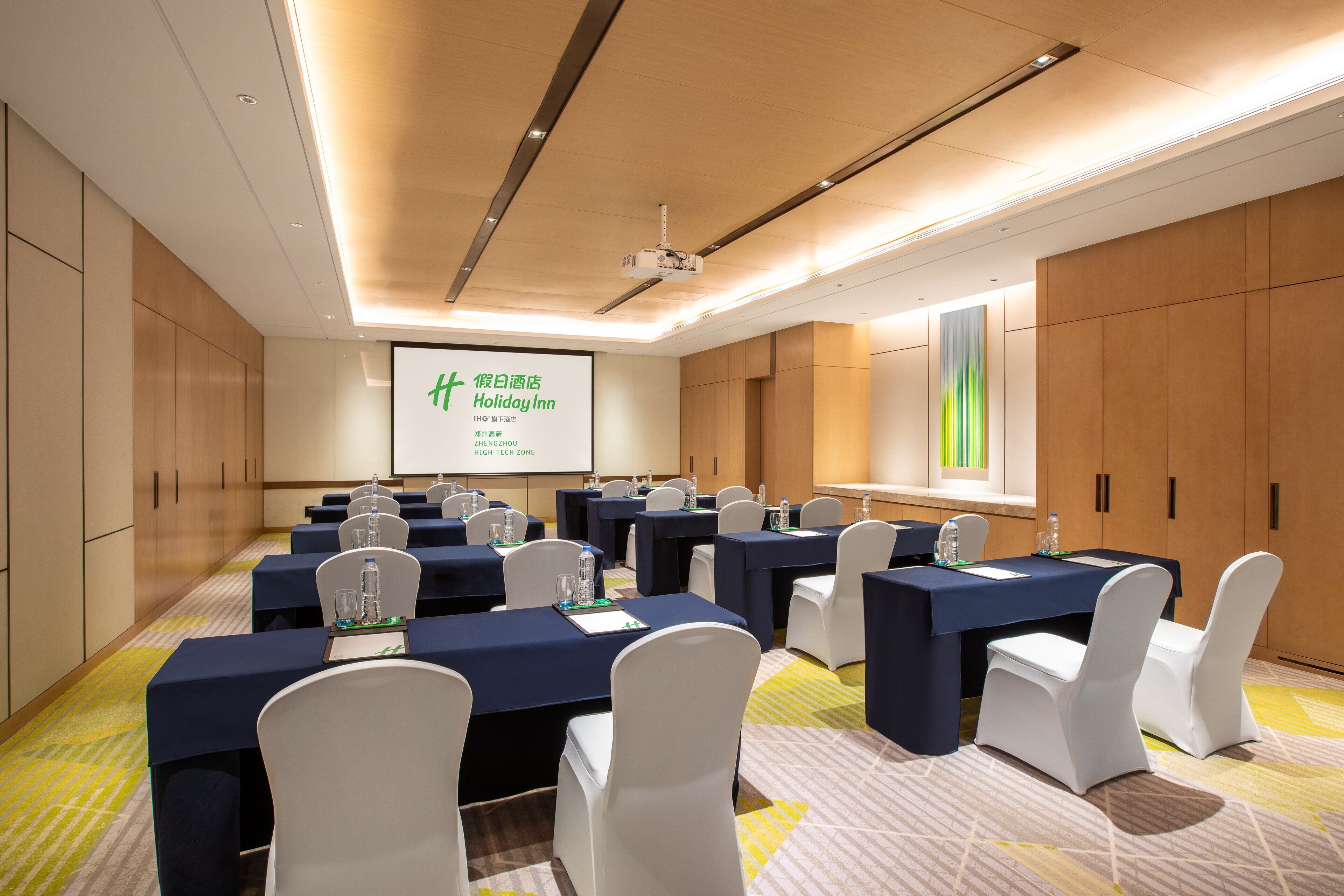 Sala de conferências com várias mesas, cadeiras brancas e uma tela de projeção exibindo uma apresentação.